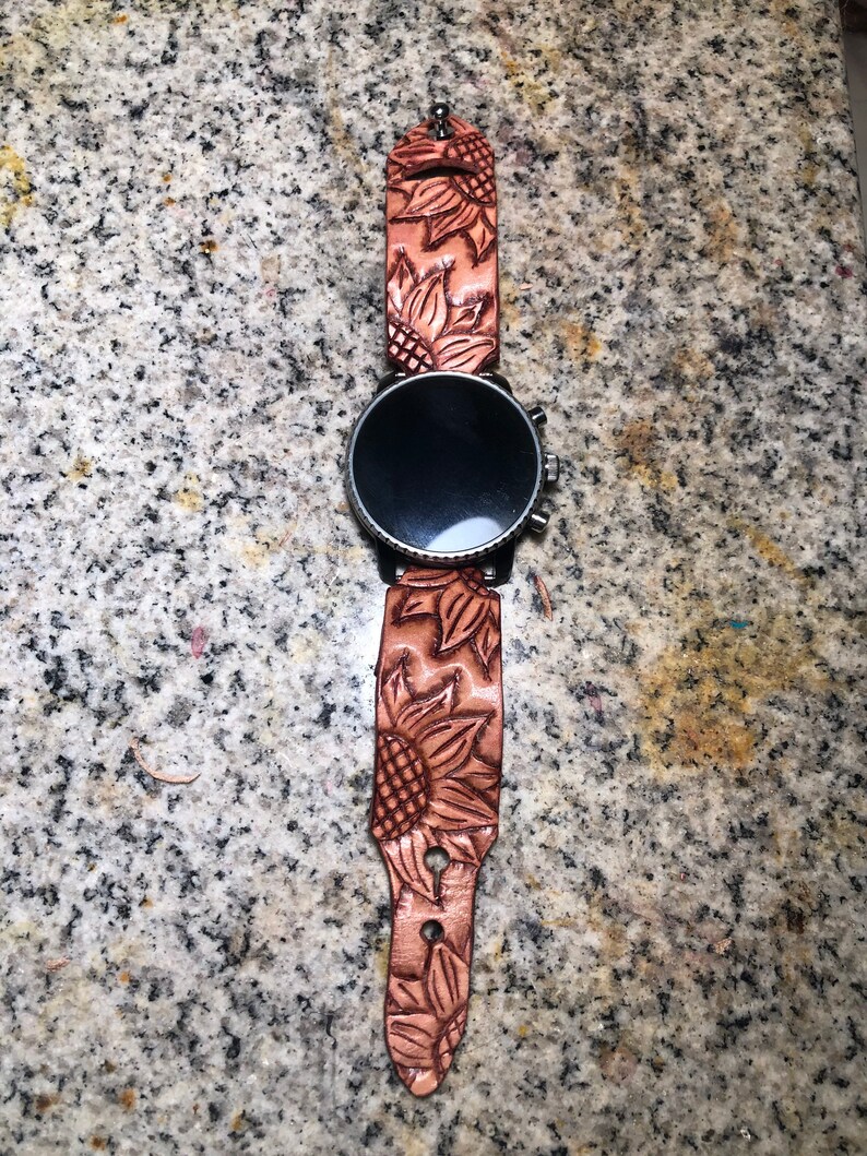 May include: A brown leather watch strap with a sunflower design. The strap is attached to a black smartwatch with a round face.