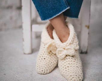 Può includere: Un paio di pantofole bianche all'uncinetto con un dettaglio a fiocco. Le pantofole sono indossate da una persona seduta su una sedia bianca.
