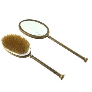 May include: A vintage gold-toned hand mirror and brush set. The mirror has a round, ornate frame and the brush has a wooden handle with a soft bristle head.