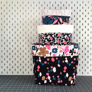 May include: A set of four fabric storage baskets with floral and geometric patterns. The baskets are stacked on top of each other, with the largest basket at the bottom and the smallest basket at the top. The baskets are all black with white, pink, and green accents.