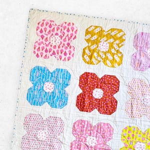 May include: A white quilt with a floral pattern. The quilt is made up of squares with different colored flowers. The flowers are pink, yellow, blue, red, and orange. The quilt is edged with a blue and white trim. The text "Lou Orth Designs" is in the bottom left corner of the quilt.