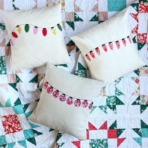 Könnte beinhalten: Drei dekorative Kissen mit weißem Hintergrund und einem Lichterketten-Design. Die Kissen liegen auf einer weißen und grünen Steppdecke mit Sternenmuster.