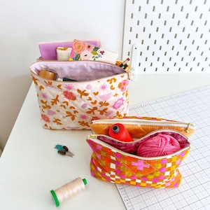 Könnte beinhalten: Zwei gesteppte Reißverschlusstaschen, eine mit Blumenmuster und die andere mit einem rosa-orangenen Karomuster. Die Taschen sind offen und mit Nähzubehör gefüllt, darunter Garn, Wolle und eine Nadel.