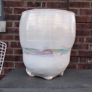 May include: A large white ceramic planter with a rounded base and a wide opening. The planter has a subtle, faded design of green, pink, and blue hues around the middle.