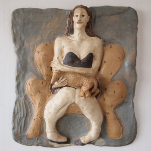May include: A ceramic wall sculpture of a woman sitting in a chair with a small animal in her lap. The woman is wearing a black top and white pants. The sculpture is painted in shades of brown, white, and black.