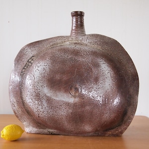 May include: A large, handcrafted ceramic bottle with a bulbous body and a narrow neck. The bottle has a textured, mottled brown and white glaze. A yellow lemon sits on the wooden surface in front of the bottle. The bottle is a decorative item.