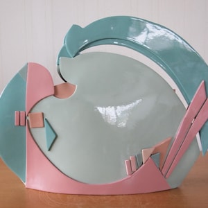 May include: A ceramic vase with a geometric design in shades of pink, teal, and beige. The vase has a unique, abstract shape and a glossy finish.