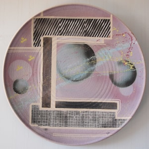 May include: A ceramic plate with a geometric design in black, white, and gray. The plate has a pink background with splashes of yellow, blue, and pink paint.