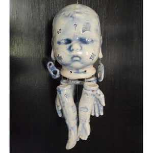 May include: A ceramic doll head with blue and white painted details, including a question mark and eyelashes. The head is attached to a string and hanging from the ceiling. The doll has two ceramic arms hanging below the head.