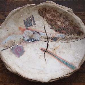 May include: A white ceramic plate with a crack running through the center. The plate has a rough, textured surface and is decorated with brown, blue, and pink accents.