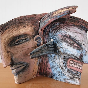 May include: A ceramic sculpture of two faces, one with a neutral expression and the other with a fierce expression. The sculpture is painted in shades of brown, blue, and pink.
