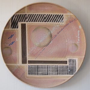 May include: A round ceramic plate with a geometric design in black, white, and blue. The plate has a light pink background with a speckled texture.