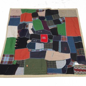 May include: A patchwork quilt featuring a mix of fabrics in green, blue, red, and black. The quilt is bordered with a beige fabric with a small repeating pattern. Some patches have initials and a red square with a design.