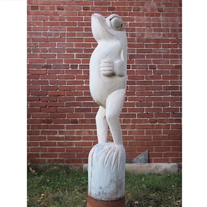 May include: A white wooden frog sculpture with a whimsical expression, standing on a round wooden base. The frog is carved with detailed features and has a textured surface.