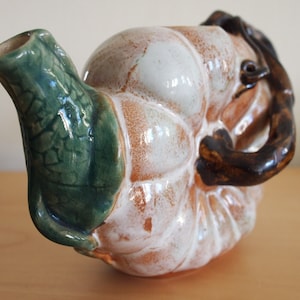 Pode incluir: Uma chaleira de cerâmica branca e marrom com um bico em forma de folha verde e uma alça marrom. A chaleira tem a forma de uma cabaça.