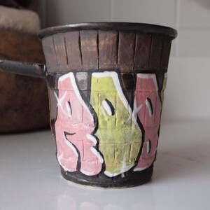 May include: Handmade ceramic mug with a dark brown, textured top and a black handle. The mug features graffiti-style lettering in pink and yellow against a black base, creating a unique, artistic design.