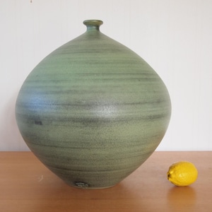 May include: A large, spherical ceramic vase with a matte, sage green finish featuring subtle horizontal bands of darker gray and olive tones. The vase has a narrow, circular opening at the top and is positioned next to a single yellow lemon.