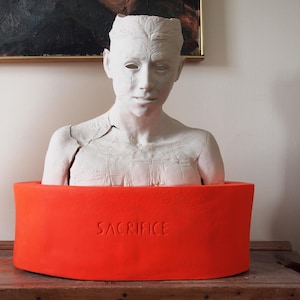 May include: A white bust sculpture of a person with a cracked chest, set within a vibrant orange cylindrical base. The word "SACRIFICE" is embossed on the base. The sculpture is displayed on a wooden surface.