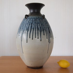 May include: A white ceramic vase with blue and black glaze, featuring a drip effect. The vase has a rounded body and two small handles on the neck.