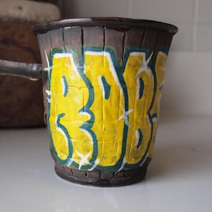 May include: Handmade ceramic mug with a dark brown exterior and a yellow graffiti-style design. The word "333" is painted in yellow with a teal outline. The mug has a small handle and is suitable for hot or cold beverages.