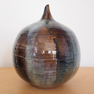 Puede incluir: Un jarrón de cerámica con cuerpo redondeado y parte superior puntiaguda. El jarrón está decorado con esmalte marrón, verde y blanco.