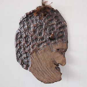 May include: A ceramic wall sculpture of a face with a textured surface and a brown feather on top.