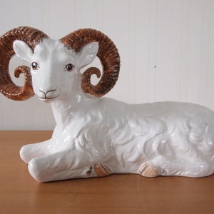 May include: White ceramic ram figurine with brown curled horns. The ram is lying down with its head turned to the side.