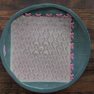 May include: A teal ceramic plate with a raised design around the edge. The plate has a pink and white patterned insert with a repeating circle design.