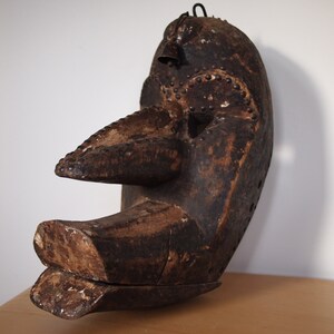 May include: A wooden mask with a long, pointed nose and a large mouth. The mask is carved with intricate details and is painted in a dark brown color. It has a metal loop on top for hanging.