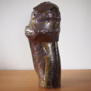 Könnte beinhalten: Eine hohe, braune Keramikvase mit einem einzigartigen, geschichteten Design. Die Vase hat eine strukturierte Oberfläche und eine leicht unregelmäßige Form.