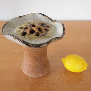 Puede incluir: Un jarrón de flores de cerámica con un esmalte moteado marrón y blanco. El jarrón tiene una parte superior ancha y festoneada con un patrón de agujeros para sujetar las flores. La base del jarrón es de forma cilíndrica.