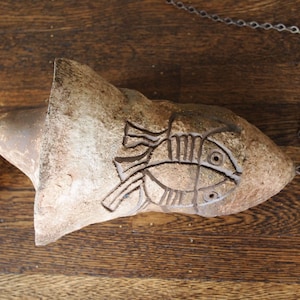 Könnte beinhalten: Eine Windspiel aus Terrakotta mit einem Fischdesign, das an einer Metallkette hängt. Das Windspiel ist braun und hat eine raue Textur.