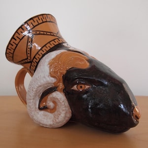 May include: A ceramic pitcher in the shape of a ram's head. The pitcher has a tan body with black and white details. The ram's head is black with tan and white accents. The rim of the pitcher has a geometric design.