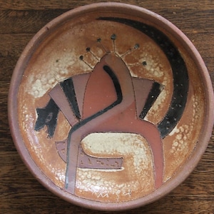 May include: A brown ceramic plate with a stylized design of a black animal with a red and black geometric pattern. The plate is signed "M.94".