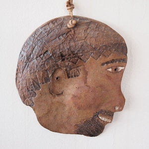 May include: A brown ceramic sculpture of a person's face in profile. The sculpture has a textured surface and is painted with brown, white, and black paint. The person has a beard and a short haircut.