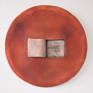 May include: A large, round, reddish-brown ceramic piece with a square cut-out. The cut-out reveals a lighter-colored square element. The piece has a textured surface and appears to be handmade. The overall design is abstract and artistic.