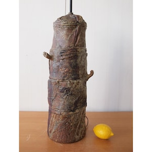 May include: A tall, cylindrical ceramic lamp base with a textured, mottled brown and earthy finish. The surface features layered, rugged clay segments and small protruding decorative elements. A bright yellow lemon sits beside the lamp base.