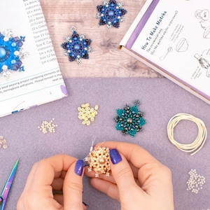 May include: A close-up of a person's hands working on a beaded snowflake craft project. The project includes a variety of beads in different colours and sizes, including iridescent blue, gold, and green. The project also includes a pair of scissors, a spool of thread, and a small bowl of beads.