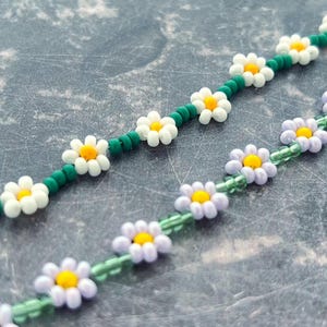 May include: Two beaded strands featuring daisy-shaped flower beads. The top strand consists of white petals with yellow centres separated by dark green spacer beads. The bottom strand features light purple petals with yellow centres and light green spacers.