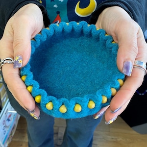 May include: A teal felt bowl with a yellow beaded trim. The bowl is being held by a person's hands.