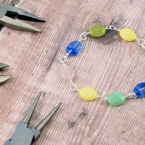 May include: A bracelet made with oval-shaped beads in shades of green, blue, yellow, and pink. The bracelet is made with silver chain links and a silver clasp. Two pairs of pliers are also in the image.