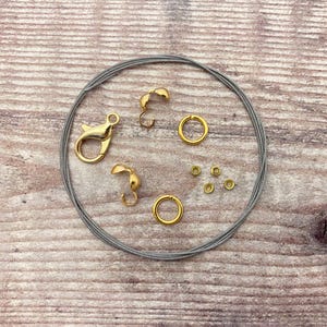 May include: A collection of jewelry-making supplies, including a gold-colored lobster clasp, crimp beads, jump rings, and cord ends. A bundle of gray beading cord is also included. The items are arranged on a light brown wooden surface.