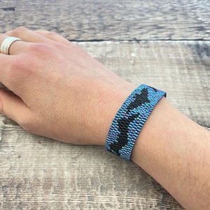 May include: A beaded bracelet in shades of blue and black is worn on a wrist. The bracelet features a geometric pattern. The background is a wooden surface. The bracelet is approximately 3.8 cm wide.