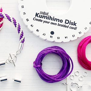Pode incluir: Um disco Kumihimo branco com texto preto que diz "mini Kumihimo Disk Create your own braided cord!" O disco tem os números 6, 7, 8, 9, 10, 11, 12, 13, 14, 15, 16, 17, 18, 19, 20, 21 e 22 ao redor da borda. Existem dois carretéis de cordão, um rosa e um roxo, e vários fechos de metal prateados.
