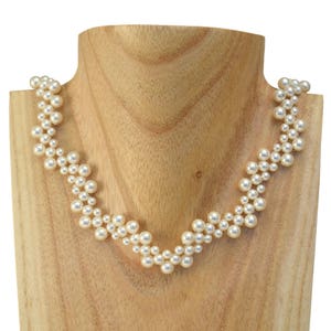 Puede incluir: Un collar de perlas blancas con un diseño en forma de V. El collar está hecho de pequeñas perlas redondas que están enhebradas juntas en un patrón de filas alternas.