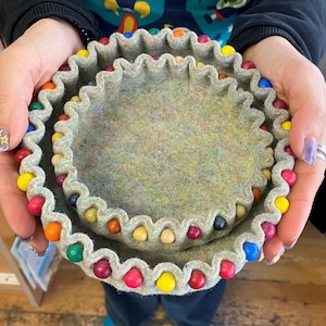 May include: Two felted bowls with scalloped edges, decorated with colorful wooden beads. The bowls are nested inside each other.