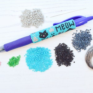 Op de afbeelding: Een blauwe pen met een kralenontwerp met een zwarte kat en het woord "MEOW". De pen is omringd door kralen in verschillende kleuren, waaronder grijs, turquoise, groen, zwart en roze. Er is ook een spoel zilverdraad.