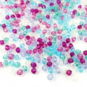 Könnte beinhalten: Eine Ansammlung facettierter Perlen in verschiedenen Farben, darunter Rosa, Lila, Türkis und Klar. Die Perlen sind verstreut und bilden eine strukturierte und farbenfrohe Darstellung. Geeignet für Schmuckherstellung und Bastelprojekte.