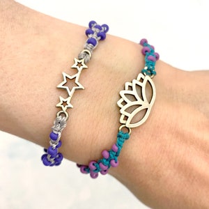 Könnte beinhalten: Zwei Freundschaftsarmbänder mit silbernen Anhängern. Ein Armband ist grau und blau mit einem Sternanhänger. Das andere Armband ist türkis und lila mit einem Lotusblütenanhänger.