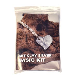 May include: A silver heart pendant necklace on a chain, resting on a piece of wood. The packaging says "Art Clay Silver Basic Kit".
